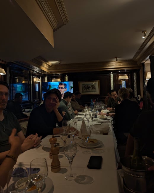 A low-light candid photograph captures a group of people seated around a long dining table in an upscale, wood-paneled restaurant, with a large television screen in the background displaying a man's face.