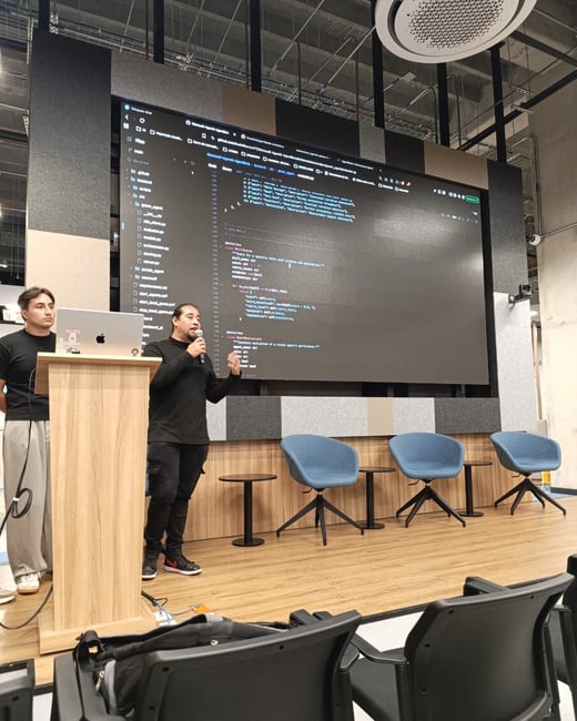 A man stands on a stage giving a presentation, gesturing with a microphone in front of a large screen displaying computer code. Another man stands nearby at a wooden podium with a laptop.