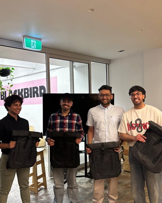 Four young men stand indoors, smiling and posing for a photo while holding dark grey roll-top backpacks, likely promotional items or prizes from an event. The background shows a large window revealing pink lettering that spells out 'BLACKBIRD'.
