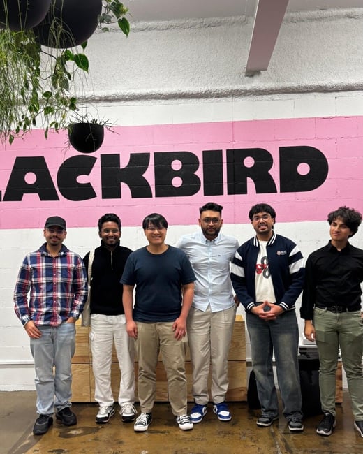 A group of six men stands side-by-side, posing for a photo indoors in front of a textured pink wall featuring large black block lettering that reads "ACKBIRD". The setting appears to be a modern or industrial office space decorated with hanging plants.
