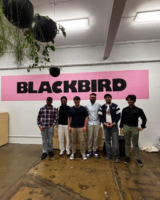 A full-length photograph of six young men posing indoors in an industrial-style space, standing in front of a large pink patch on a white wall featuring the black text "BLACKBIRD."