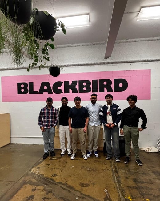 A group portrait featuring six young men standing in a line indoors against a white wall that has the word "BLACKBIRD" painted in black capital letters on a large pink rectangular background. The setting appears to be a casual or industrial space with a rough concrete floor and hanging plants.