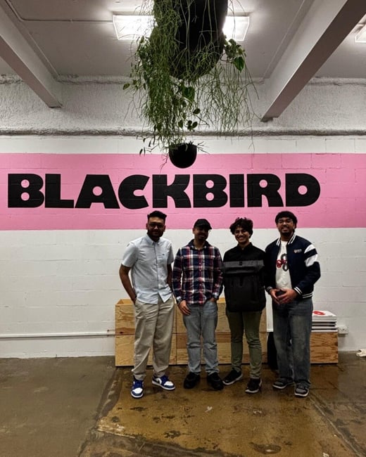 Four young men are posing together for a group photo indoors, standing in front of a white wall featuring a bright pink horizontal stripe with the word "BLACKBIRD" painted in large black capital letters.
