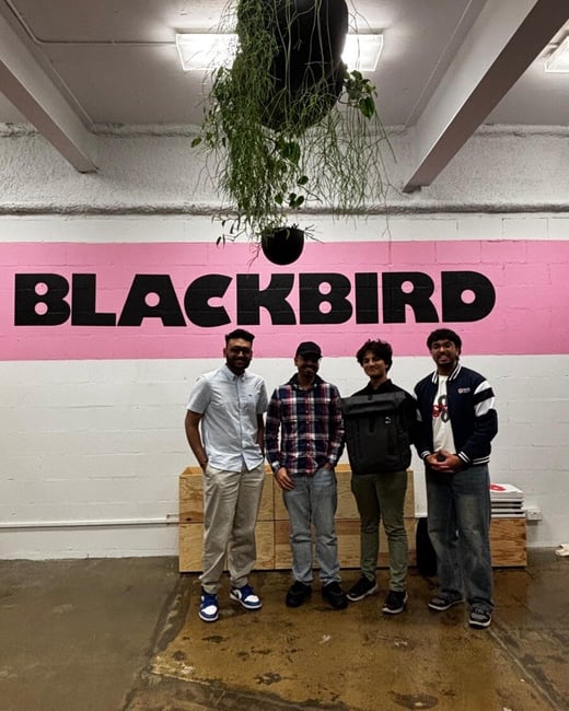 A group of four men stands posing for a photo in an indoor setting, centered against a white brick wall featuring a wide pink stripe with the bold black text 'BLACKBIRD'. A large black planter with trailing green vines hangs from the ceiling above the text.