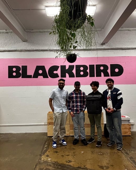 A group photo of four young men standing indoors in front of a white brick wall featuring a large horizontal pink stripe with the word "BLACKBIRD" painted in black, bold lettering.