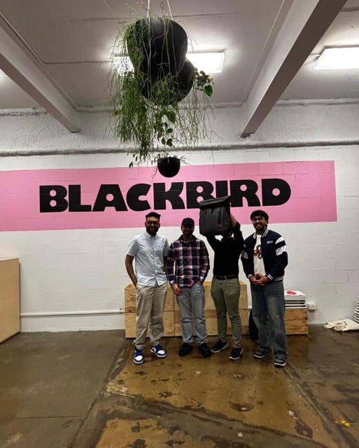 A group photo of four men standing indoors in a large, industrial-style room in front of a pink wall section displaying the word "BLACKBIRD" in large black capital letters.