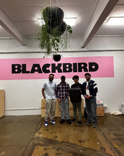 Four young men pose for a group photo indoors in front of a large pink rectangular mural featuring the word "BLACKBIRD" in bold black capital letters.