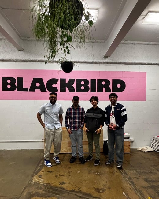 A group photo of four men standing indoors in front of a white brick wall featuring a large pink stripe with the black text "BLACKBIRD" painted on it. Hanging plants are visible from the ceiling above the subjects.