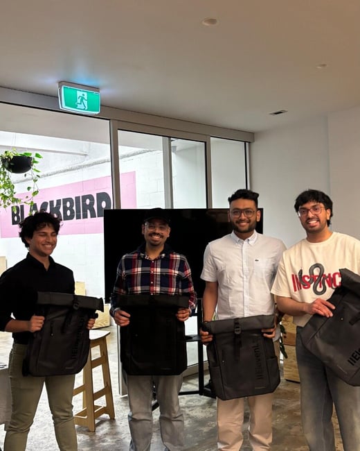 A photograph showing four young men standing indoors, smiling at the camera while holding identical black roll-top backpacks. The background features a large black 'BLACKBIRD' sign against a pink stripe in what appears to be a modern office or event space.
