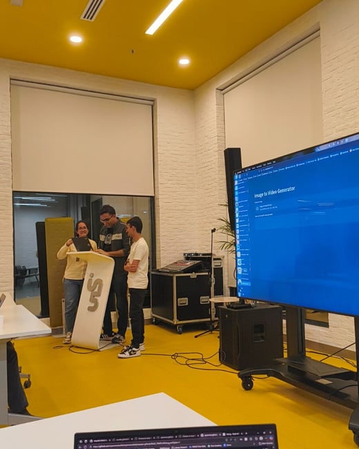 A candid photograph taken indoors during a technical presentation or discussion, showing three people gathered around a podium while a large screen displays the title "Image to Video Generator."