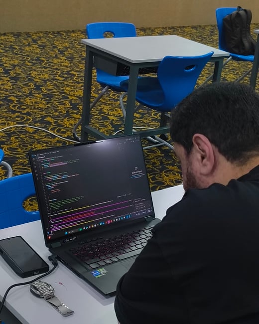 A man is seen from behind, sitting at a white desk and working on a laptop that displays lines of code in a dark-themed editor. He is in a room with blue plastic chairs and a distinctive gold and black patterned carpet.