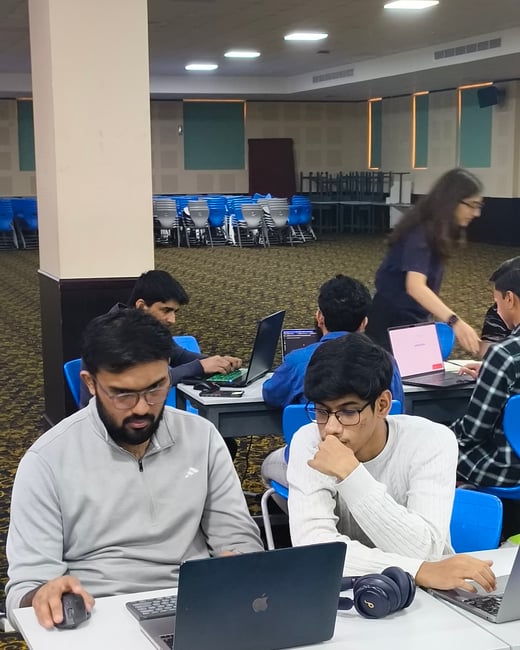 A group of young people are working on laptops at tables in a large indoor hall, with two men in the foreground focused on a shared screen.