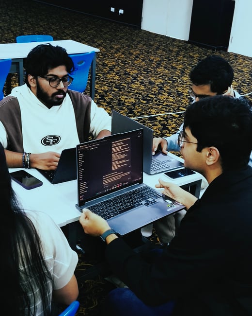 A group of young adults are gathered around a table in an indoor setting, focused on their laptops and collaborating on a project.