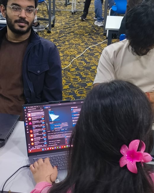 A young woman with a pink flower hair clip is working on a laptop showing code, while a man in glasses sits across from her in a large room with patterned carpet.