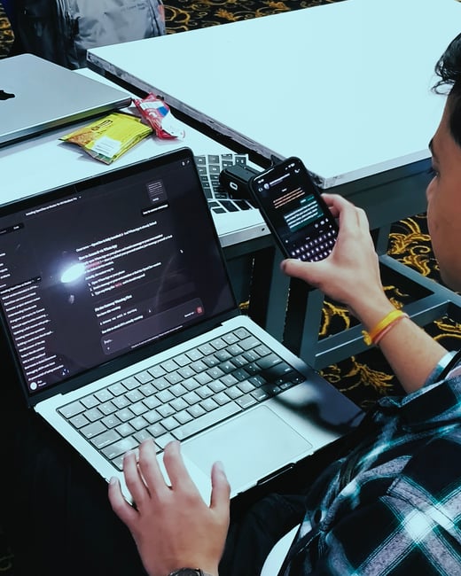 A person is sitting at a white table, using a laptop and a smartphone simultaneously while following a technical tutorial.