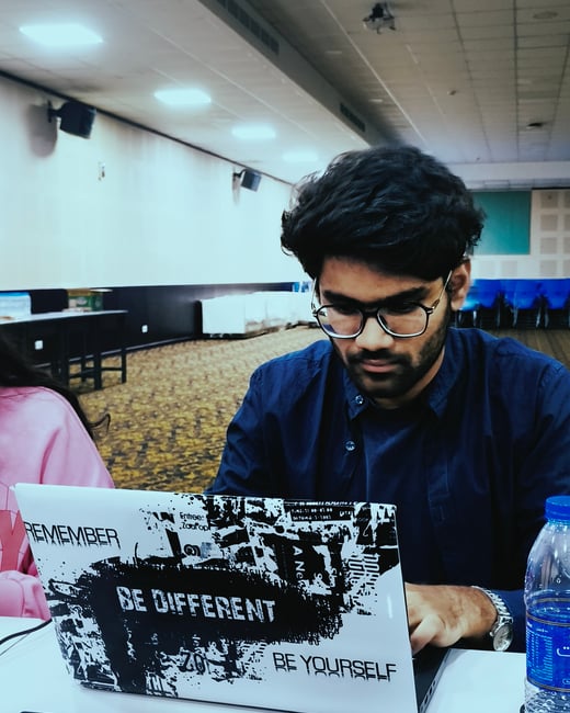 A young man with glasses and dark hair is focused on working on a laptop in a large indoor hall. The laptop's lid is covered with a black and white sticker featuring motivational phrases.
