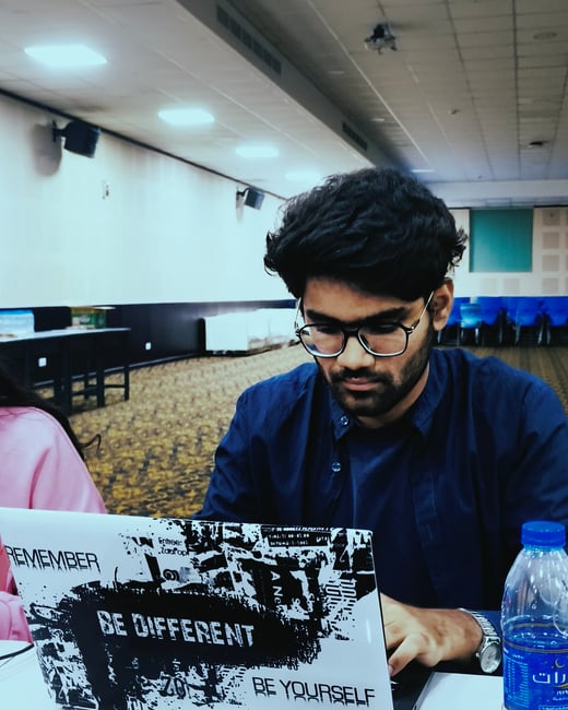 A young man with glasses and a beard is sitting at a table, focused on his laptop in a large indoor hall. The laptop is decorated with stickers that read 'REMEMBER', 'BE DIFFERENT', and 'BE YOURSELF'.