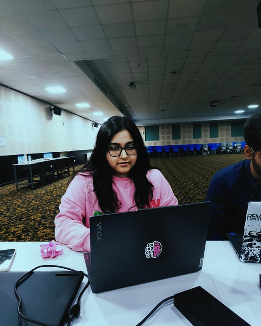 A young woman wearing glasses and a pink sweatshirt is focused on her laptop in a large, dimly lit hall.
