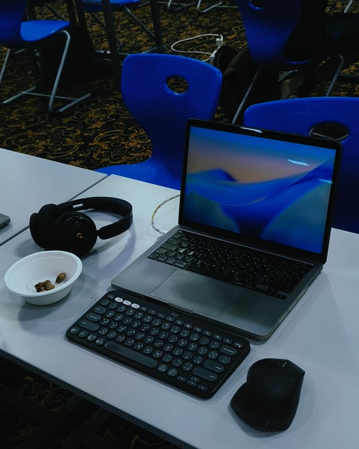 A laptop setup on a white table featuring an external keyboard, mouse, headphones, and a small bowl of snacks, with blue chairs in the background.