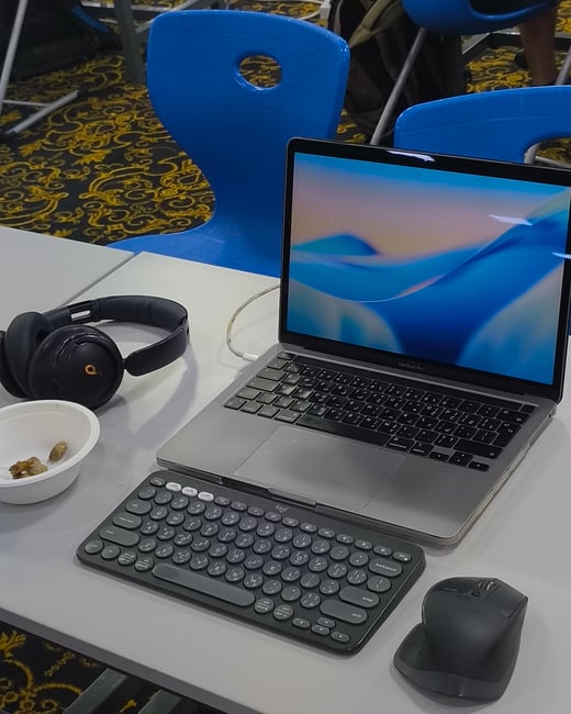 A workspace setup on a white table featuring a laptop, an external wireless keyboard, a mouse, and headphones, located in a room with blue chairs and a patterned carpet.