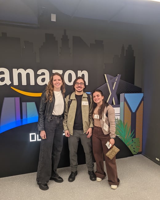 Three people stand posing together in front of a black backdrop featuring the Amazon logo and city silhouettes.