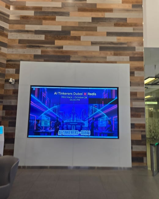 A photograph of an indoor lobby or hallway featuring a large digital display screen mounted on a white panel below a wall decorated with horizontal wood planks. The screen displays promotional graphics and text for an event titled 'AI Tinkerers Dubai X Redis Mini Hack'.