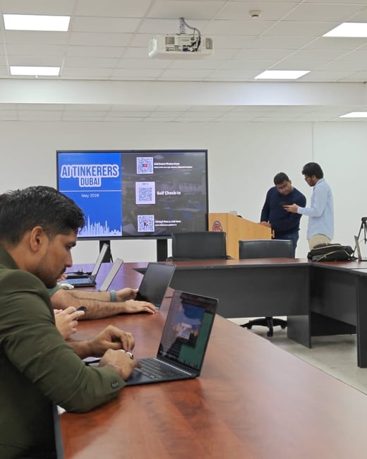 A photograph showing an indoor conference room during an AI Tinkerers Dubai event, with participants sitting at a wooden table using laptops while others stand near a presentation screen in the background.