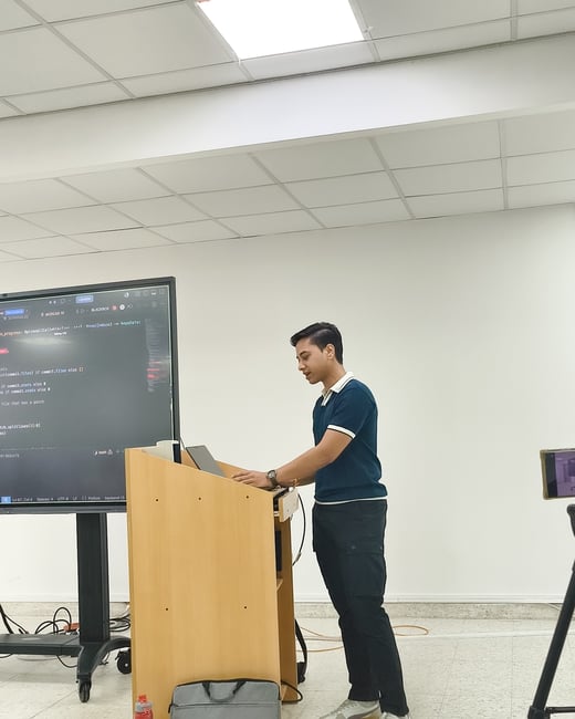 A young man stands at a wooden podium, using a laptop in front of a large display screen showing Python code in what appears to be a classroom or presentation room.
