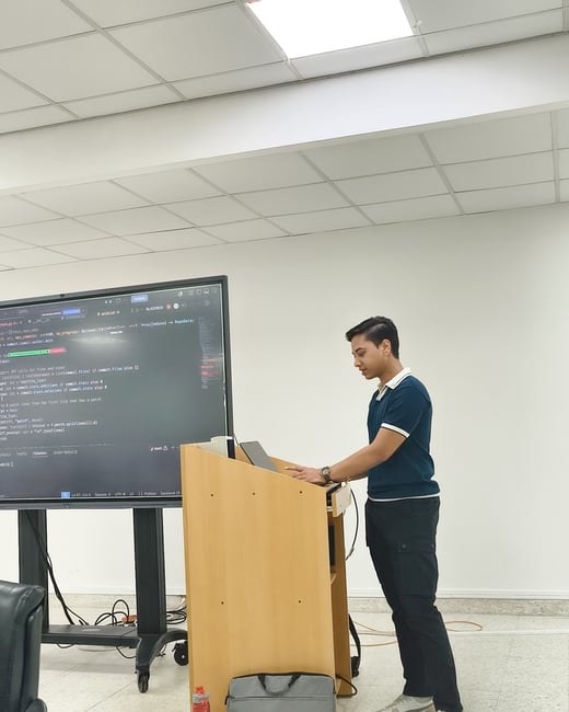 A young man stands at a wooden podium, interacting with a laptop that is connected to a large screen displaying Python source code.
