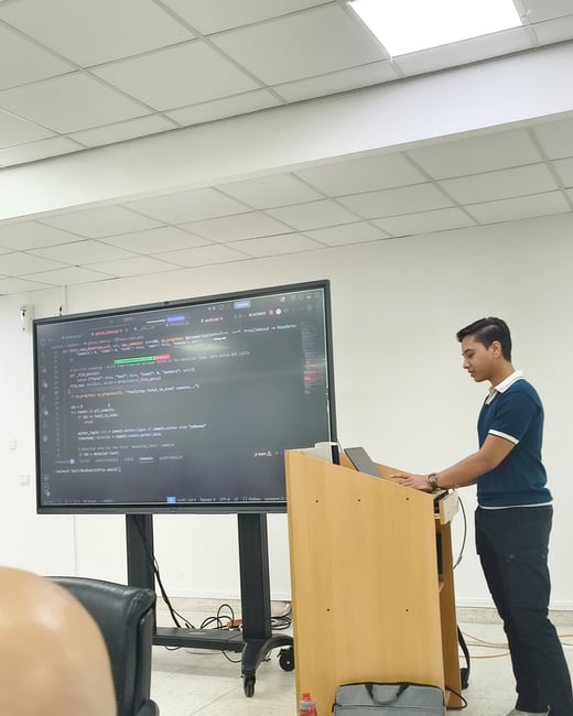 A young man stands at a wooden podium, typing on a laptop during a presentation. Behind him, a large screen displays Python source code in a dark-themed text editor.
