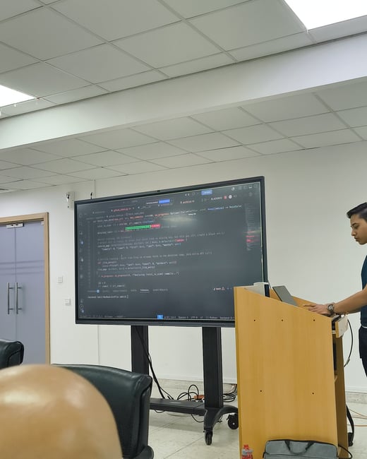 A photograph showing a young man presenting at a wooden podium, using a laptop connected to a large screen displaying Python code in a classroom or office setting.