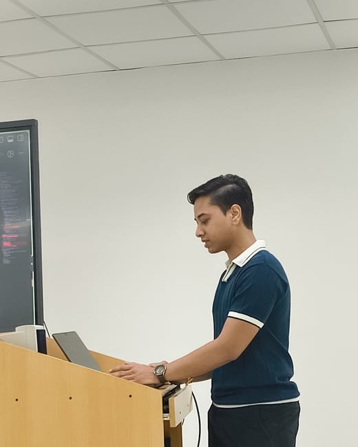 A young man stands in profile at a wooden lectern, using a laptop during a presentation or lecture in a brightly lit room.