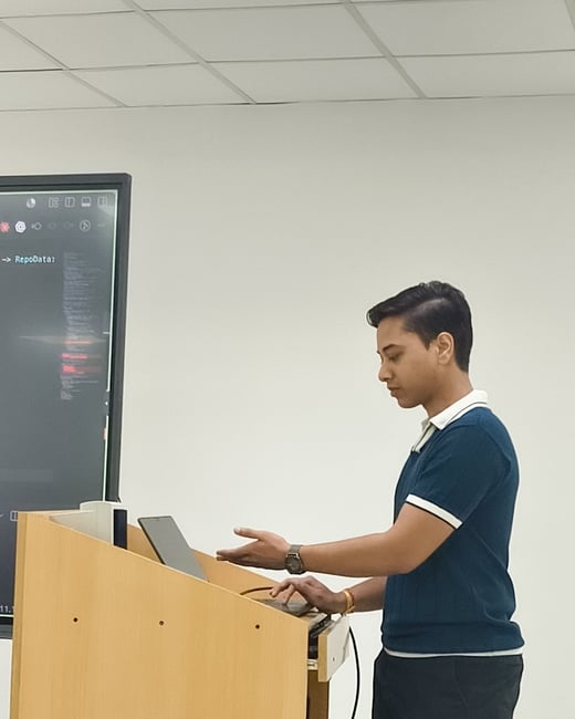 A young man stands in profile at a wooden podium, gesturing towards a laptop while presenting in front of a large screen displaying code.