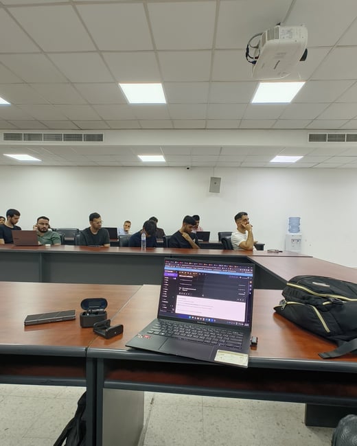 An indoor classroom or lecture hall setting showing a laptop open on a wooden desk in the foreground, with a group of people sitting at desks in the background listening to a presentation.