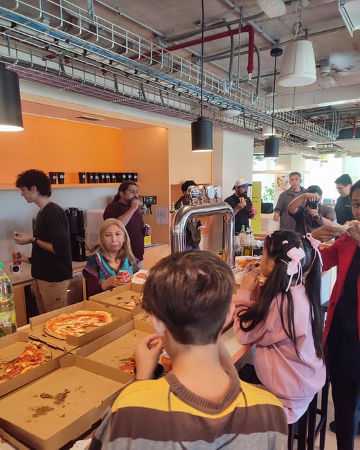 A group of people, including children and adults, are gathered in a modern office kitchen area eating pizza from several open boxes.