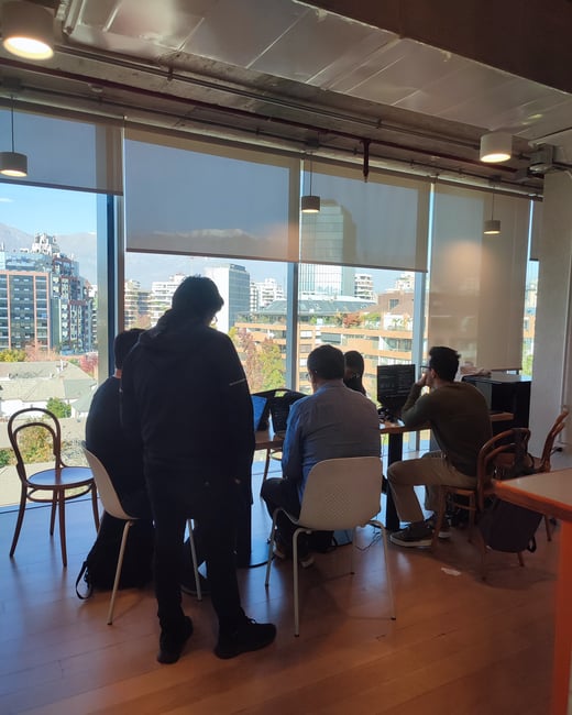 A group of people are gathered around a table with laptops in a modern office space, featuring large windows with a view of a city skyline.