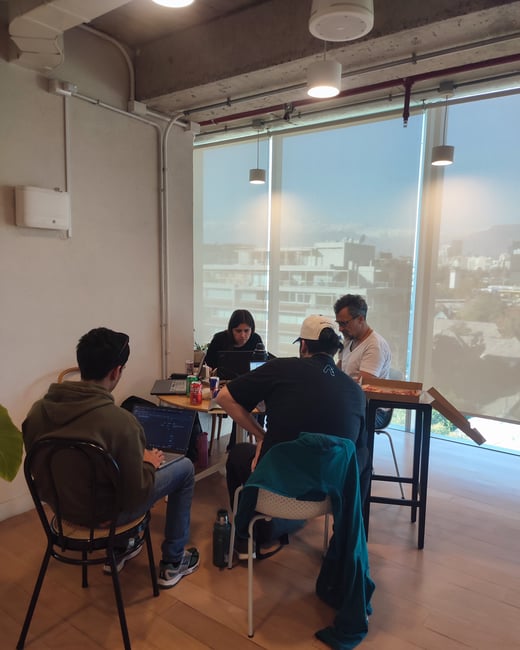 A group of four people are gathered around a table in a modern office space, working on laptops with a large window overlooking a city in the background.