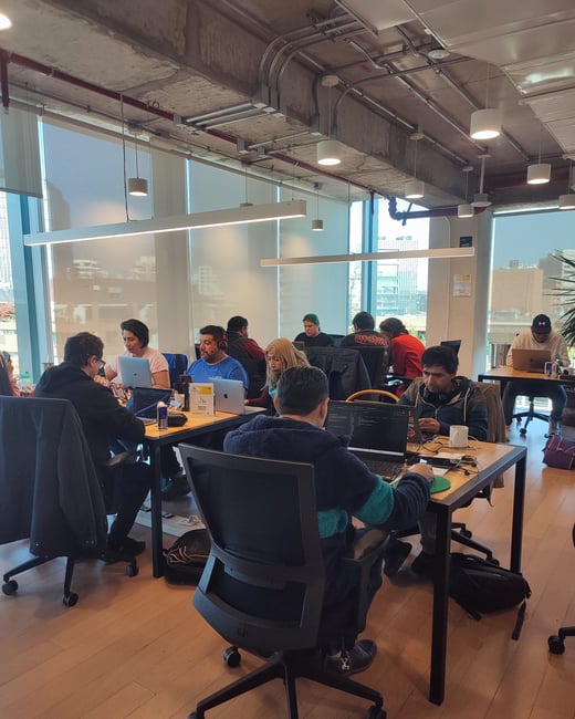 A group of people are working on their laptops in a modern, industrial-style office or co-working space with large windows.