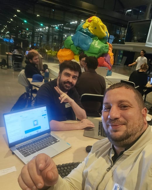 Two men pose for a close-up selfie while sitting at a table with laptops in a large, dimly lit indoor space, likely during a hackathon or tech event. A large, brightly colored, abstract sculpture dominates the background.