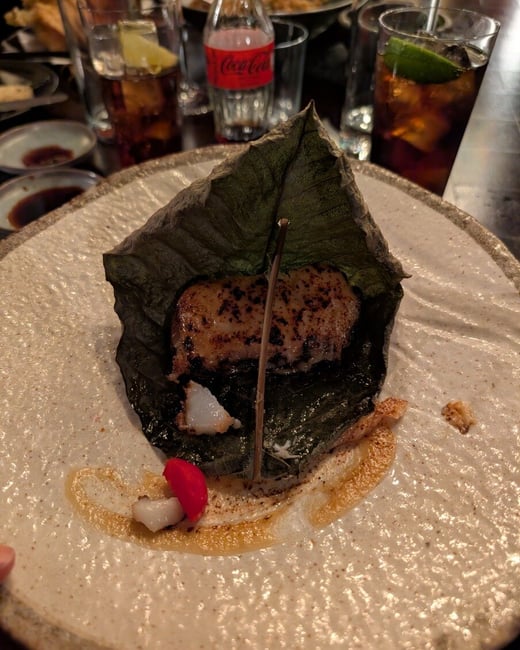 A close-up photograph of a gourmet dish served on a large leaf atop a textured ceramic plate, with drinks and a Coca-Cola bottle in the background.