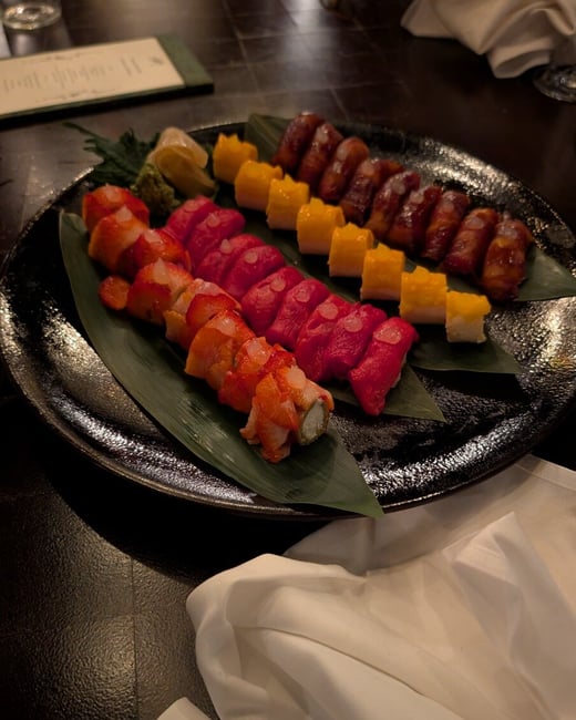 A close-up photograph of a variety of colorful sushi rolls arranged on a black platter with banana leaves in a restaurant setting.