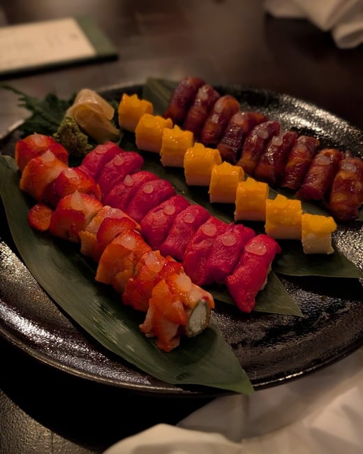 A close-up photograph of a vibrant sushi platter featuring four distinct rows of colorful sushi pieces arranged on large green leaves over a textured black plate.