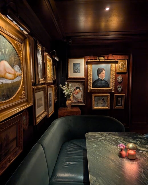 A moody, dimly lit interior of a high-end restaurant or lounge, featuring a dark green velvet booth and a marble table set against a wall covered in ornate framed paintings.