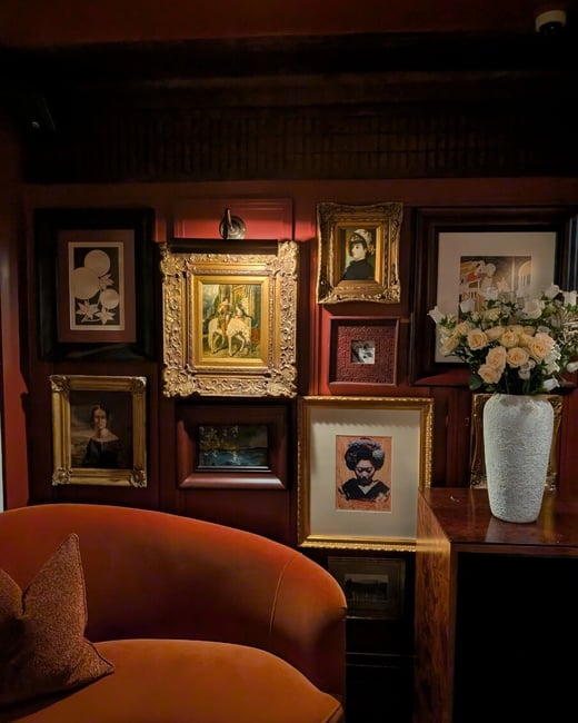 A warm, dimly lit interior featuring a gallery wall of various framed artworks above a curved orange velvet sofa and a wooden cabinet with a white vase of roses.