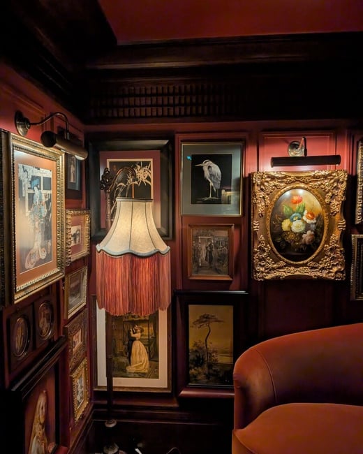 A moody and atmospheric interior corner featuring a gallery wall of various framed artworks and a vintage floor lamp with a long fringed shade.