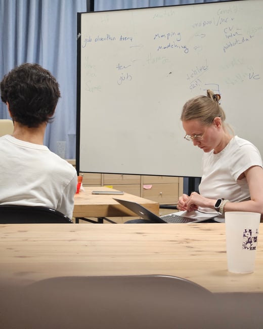 A candid indoor photograph capturing two people seated at a wooden table during a work session or meeting, with one person using a laptop and a whiteboard covered in handwritten notes visible in the background.