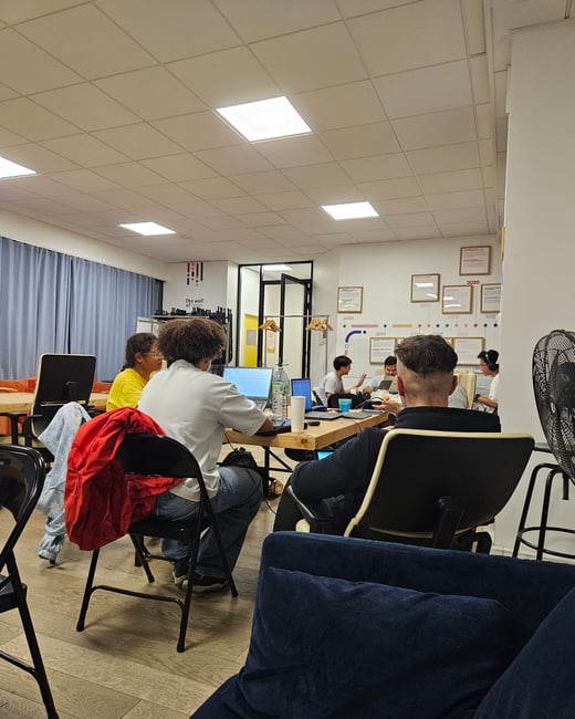 A candid photograph showing a diverse group of young adults working or collaborating around a wooden table in a brightly lit indoor office or co-working space.
