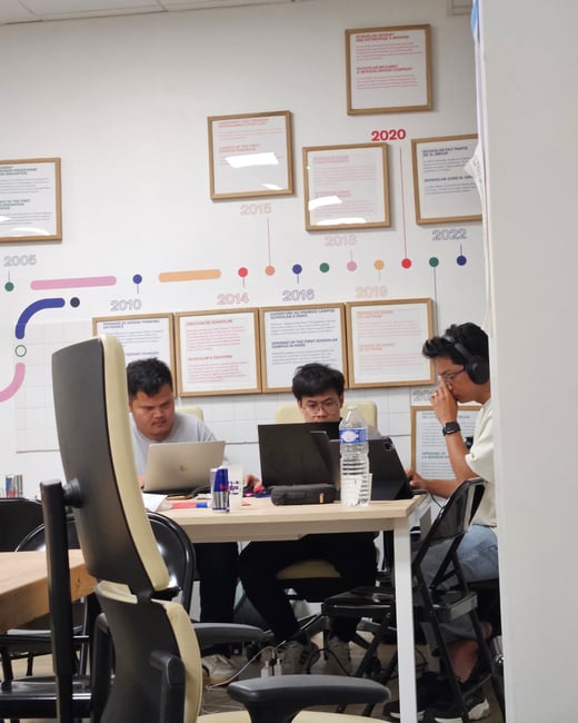 Three young men are seated at a table working on laptops and tablets in an indoor office or co-working space. The background features a wall timeline graphic detailing the history of an organization called "Schoolab" with key dates and framed descriptions.