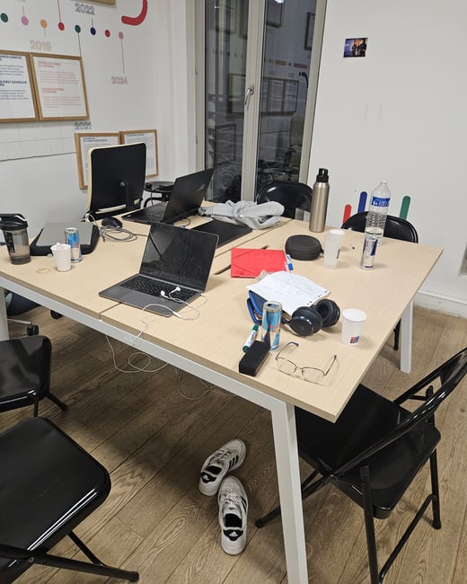 A candid indoor photograph capturing a cluttered office or study table featuring multiple closed and open laptops, various drinks (Red Bull, water), headphones, and personal items, with a pair of white sneakers resting underneath. The background wall displays a colorful timeline graphic and framed documents.
