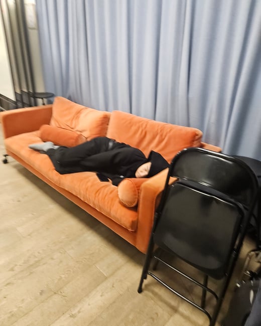 A candid indoor photo showing a person resting or sleeping on a long, bright orange velvet sofa, partially covered by black clothing.